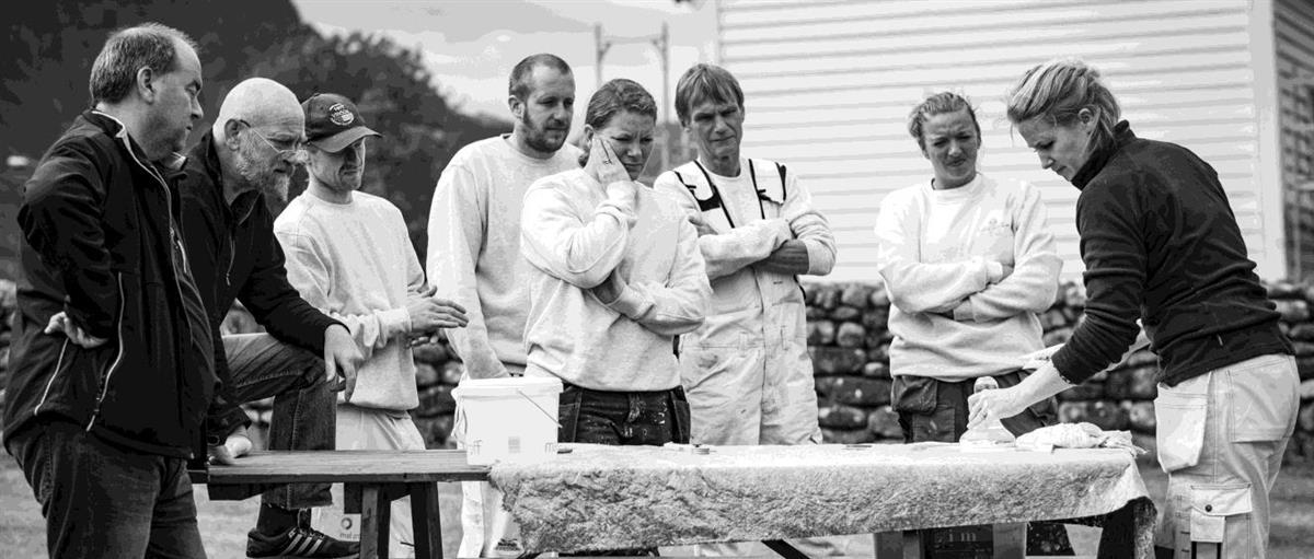 Flere menn og kvinner står rundt et bord. De er ute. På bordet er det malingspann og et tøystoff. I bakgrunnen er en steinmur og et trekledd bygg. Fotografi. 