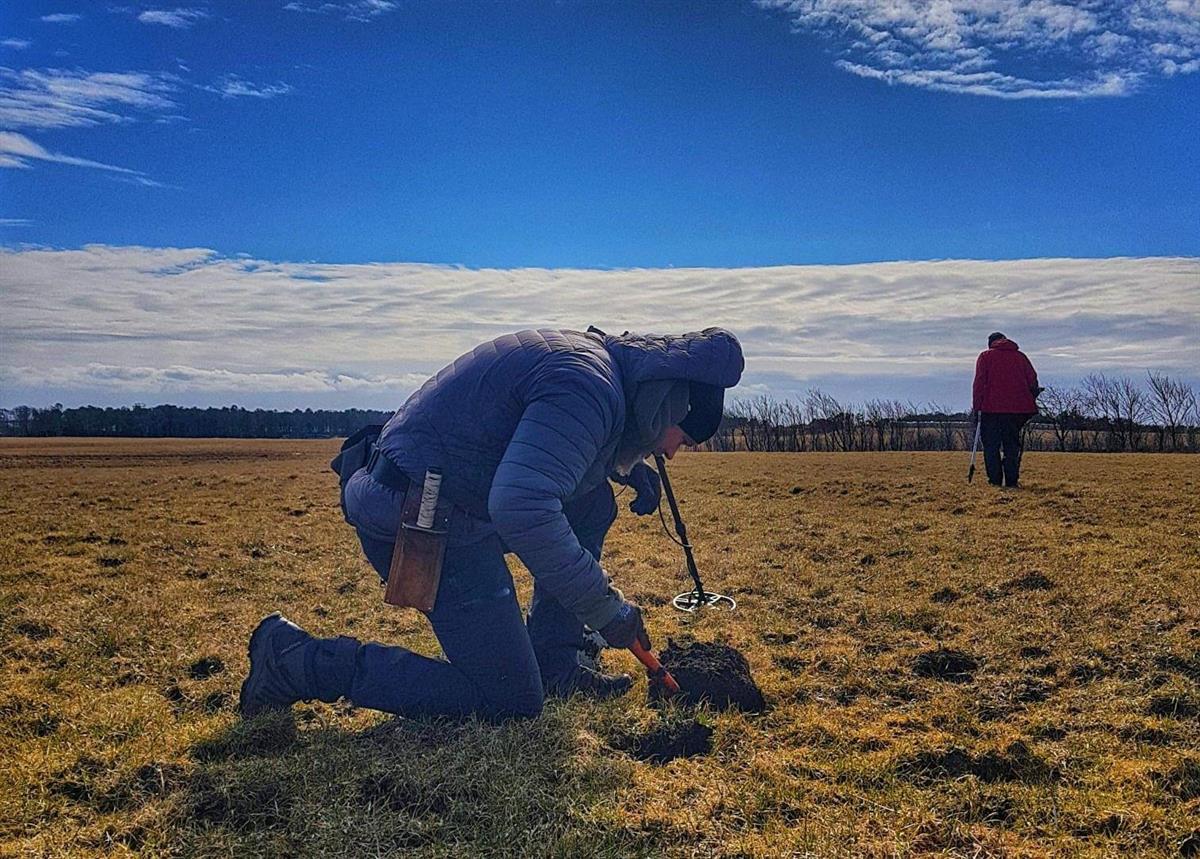 To personer på et jorde. Ene personen står oppreist og går. Andre personen sitter på kne og ser ned i et lite hull i jorden. Fotografi