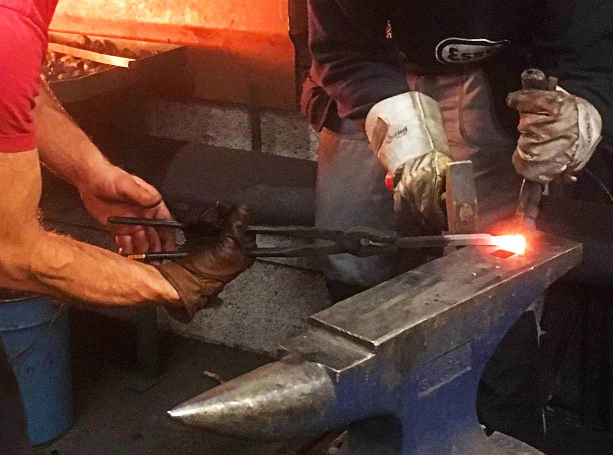 En glødende jernstang holdes mot en armbolt. Det kan skimtes to menn som arbeider med jernstangen. I bakgrunnen ser vi essen. Fotografi